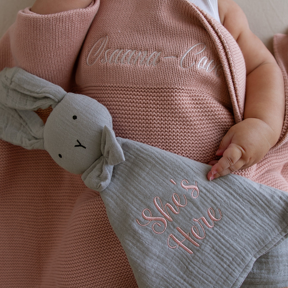 Close-up of a stone grey organic cotton muslin bunny baby comforter with knotted corners and a cute bunny head with a bow tie, partially covering a pink blanket with embroidered text.