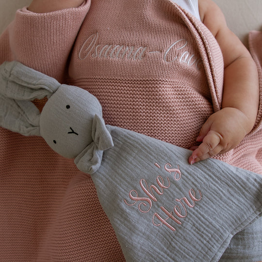 Close-up of a stone grey organic cotton muslin bunny baby comforter with knotted corners and a cute bunny head with a bow tie, partially covering a pink blanket with embroidered text.